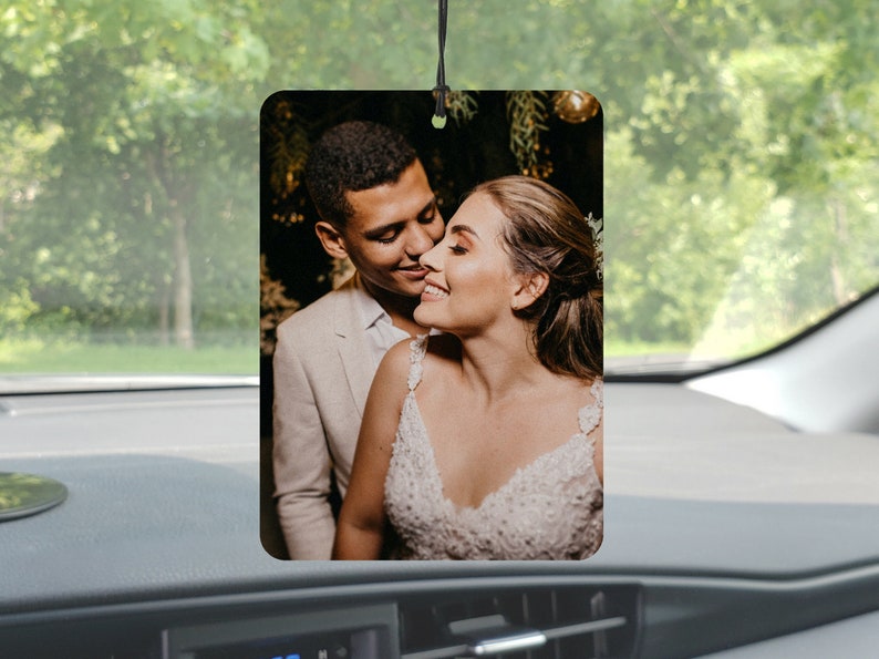 May include: Rectangular car air freshener featuring a romantic photo of a smiling couple. The image shows a man in a light-colored suit and a woman in a white wedding dress. The air freshener hangs from a black string.