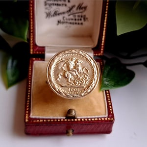 May include: A gold-colored ring with a coin-like design featuring a rider on a horse, dated 2001. The ring is set in a red jewelry box with a cream-colored interior, and the box is open. The ring is the focal point of the image.