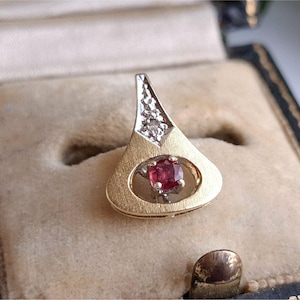 May include: A gold pendant with a teardrop shape, featuring a small, square, red gemstone at the center. The top portion has a diamond-studded design. The pendant is displayed inside a vintage jewelry box with a beige interior.