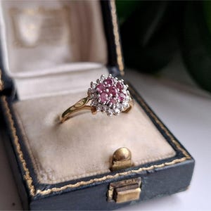May include: A gold ring featuring a cluster of round pink gemstones at the centre, surrounded by a halo of small clear white stones. The ring has a slender gold band and is presented inside a dark blue velvet-lined jewellery box.