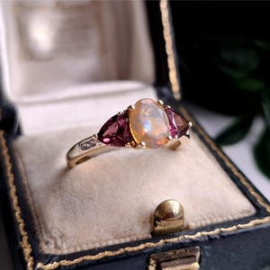 Könnte beinhalten: Ein Goldring mit einem zentralen, oval geschliffenen, irisierenden Opal, der von zwei dreieckig geschliffenen, dunkelrosa Edelsteinen flankiert wird. Der Ring sitzt auf einer polierten Goldschiene und zeigt ein klassisches Drei-Stein-Design.