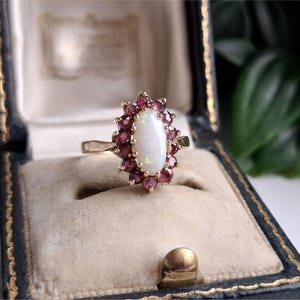 May include: A gold ring featuring a central oval-shaped white opal cabochon surrounded by a halo of small, round, deep red garnet gemstones. The ring is set in a yellow gold band, showcasing a cluster design with a polished metal finish.