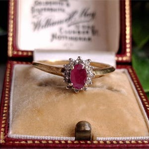 Könnte beinhalten: Goldener Ring mit einem ovalen Rubin-Edelstein, umgeben von kleinen Diamanten. Der Ring wird in einer rot-goldenen Schmuckschatulle mit cremefarbenem Innenraum präsentiert.