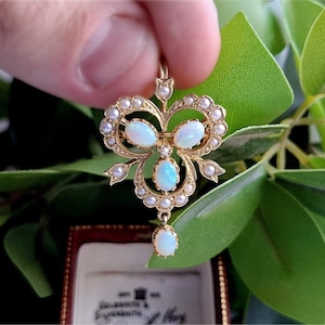 May include: A gold-toned pendant with opal and pearl accents. The pendant features three oval opals and several smaller pearls, arranged in a bow-like design. The pendant is held against a backdrop of green leaves.