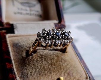 May include: A gold ring with three black gemstones surrounded by smaller white gemstones. The ring is in a brown velvet ring box.