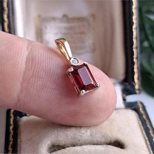 Pode incluir: Um pingente com uma pedra preciosa retangular de cor vermelho-escura em lapidação esmeralda, montada num engaste de metal prateado. Uma pequena pedra clara redonda está posicionada acima, ligada a uma argola de metal dourado.