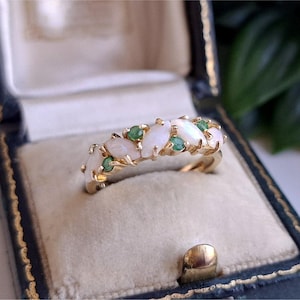 May include: A gold ring featuring a row of marquise-cut white opals alternating with small round green emeralds. The stones are set in a yellow gold band, creating a delicate and detailed design that showcases the contrast between the gems.