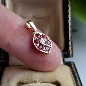 Può includere: Un pendente in oro dal design a forma di foglia, con un quadrato centrale di pietre bianche circondato da pietre preziose rosse. Il pendente è tenuto da un dito, mettendo in risalto i suoi dettagli e la sua maestria.