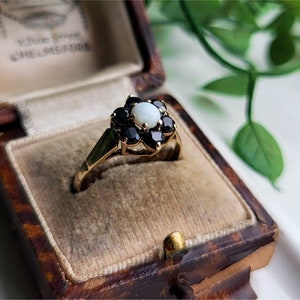 May include: A gold ring with a white opal center stone surrounded by black gemstones. The ring is in a brown velvet ring box.