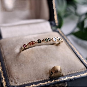 May include: A thin gold band ring featuring a series of small, multi-colored gemstones set in alternating round and marquise-shaped bezels. The stones include shades of pink, orange, green, and blue, creating a vibrant, delicate arrangement.