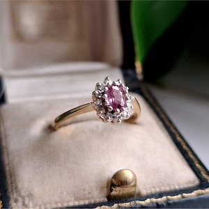 May include: A gold ring featuring an oval-shaped pink gemstone at the centre, surrounded by a halo of small, clear, round-cut stones. The band is polished gold, and the setting for the stones is silver-toned metal, creating a classic cluster design.