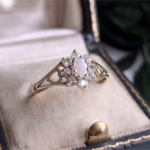 May include: A gold ring featuring an oval-shaped opal center stone surrounded by a cluster of small, clear, round-cut stones. The band has a decorative openwork design with heart-shaped cutouts on each side of the central stone setting.