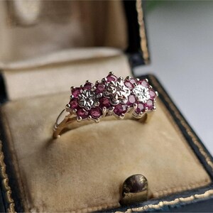 May include: A gold ring with three floral clusters. Each cluster features a central diamond surrounded by smaller, vibrant red gemstones. The ring is displayed in a vintage, velvet-lined jewelry box with a dark blue exterior.