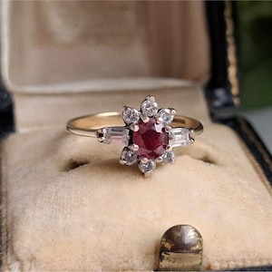 May include: A gold ring with a central oval ruby gemstone, surrounded by small diamonds and baguette-cut diamonds. The ring is set in a vintage-style setting and is displayed in a jewelry box.