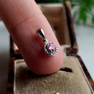 May include: A small pendant with a pink gemstone surrounded by a halo of smaller white gemstones. The pendant is made of silver and has a simple bail.