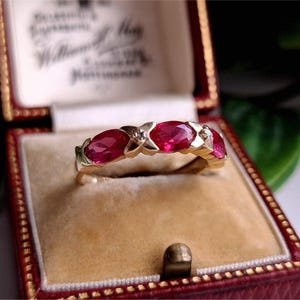 Könnte beinhalten: Ein goldener Ring mit fünf oval geschliffenen, leuchtend roten Edelsteinen. Der Ring ist in einem goldenen Band mit kleinen Diamantakzenten gefasst. Der Ring wird in einer roten Vintage-Schmuckschatulle präsentiert.