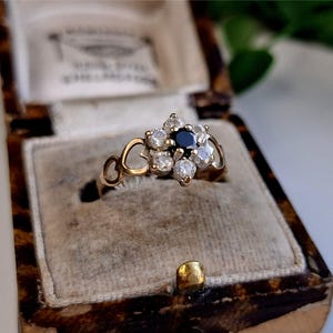 Op de afbeelding: Een vintage gouden ring met een bloemmotief. De ring heeft een donkerblauwe middensteen omringd door heldere, ronde accentstenen. De ring is gezet in een gouden band met hartvormige details. De ring wordt gepresenteerd in een vintage sieradendoos.