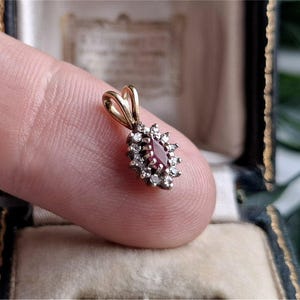 May include: A gold pendant featuring a central marquise-cut red gemstone surrounded by a halo of small round white stones. The pendant has a gold bail at the top for attachment to a chain, showcasing a classic cluster design with metallic tones.
