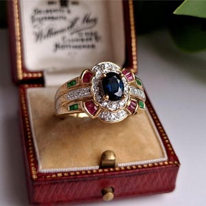 May include: A gold ring with a large oval sapphire center stone, surrounded by diamonds and smaller red and green gemstones. The ring is displayed in a red jewelry box with a gold clasp and a cream-colored interior.