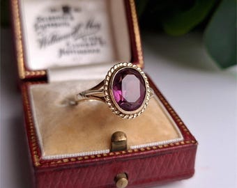 Puede incluir: Un anillo de oro con una piedra preciosa ovalada de color púrpura oscuro. La piedra preciosa está engastada en un marco dorado con un diseño de cuerda. El anillo se muestra en un joyero rojo.