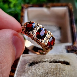 May include: A gold ring featuring three oval-shaped red gemstones and small white gemstones set in a gold band.