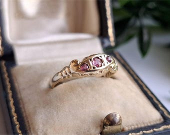 May include: A gold ring with three pink gemstones set in a decorative band. The ring is displayed inside a blue and gold jewelry box with a beige interior. The ring's design features intricate details and a vintage aesthetic.