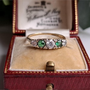 Puede incluir: Un anillo de oro con cinco piedras preciosas, incluyendo piedras claras, verdes y rosas, descansa en un joyero rojo con interior crema. El anillo está centrado, mostrando su diseño y el contraste de las piedras.