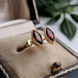 May include: A pair of stud earrings featuring marquise-cut deep red garnet gemstones set in polished yellow gold bezels. The earrings are displayed in a vintage-style jewelry box with a beige velvet interior and a dark blue exterior frame.