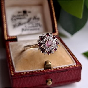 May include: A gold ring with a cluster of gemstones. The ring features a central pink gemstone surrounded by smaller white and dark red stones. The ring is displayed in a red jewelry box with a cream-colored interior.