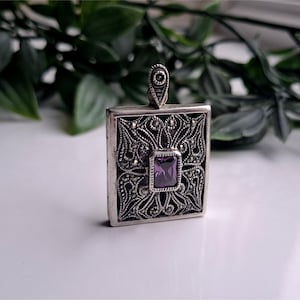Peut inclure: Un pendentif carré argenté avec des détails complexes en filigrane et en marcassite. Le pendentif présente une pierre précieuse violette carrée au centre. La bélière est une petite boucle décorative. Fond blanc avec feuillage vert.
