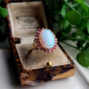 May include: A gold ring featuring a large oval cabochon opal centre stone surrounded by a halo of small, round, deep pink rubies. The ring is displayed inside a vintage-style brown patterned jewellery box with a beige velvet interior lining.