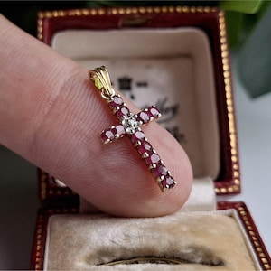 May include: Gold cross pendant with red gemstones and a central diamond cluster. The cross is held in a vintage red jewelry box with a cream-colored interior. The pendant is a religious symbol.