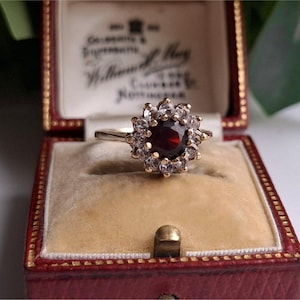 Könnte beinhalten: Ein goldener Ring mit einem großen, runden, dunkelroten Edelstein in der Mitte, umgeben von kleineren, klaren Edelsteinen in einem floralen Muster. Der Ring wird in einer roten Vintage-Schmuckschatulle mit cremefarbenem Innenraum präsentiert.