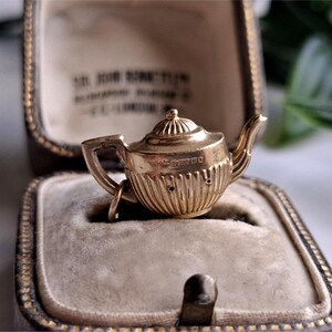 May include: A miniature gold teapot charm with a ribbed design, displayed in a vintage jewellery box. The teapot features a handle, spout, and lid. The box is brown with a cream-coloured interior.