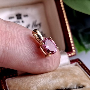 May include: A gold pendant featuring a pink oval gemstone. The pendant is held by a finger, highlighting the setting's details and the gemstone's colour. The pendant is in a velvet lined box.
