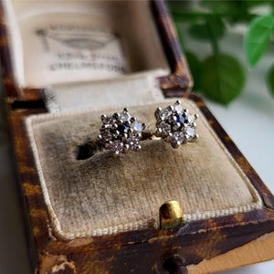 Puede incluir: Un par de pendientes de plata con un racimo de pequeñas piedras preciosas blancas y azules en forma de flor. Los pendientes están en una caja de joyería de terciopelo marrón.