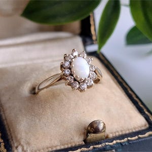 May include: A gold ring with a central oval opal stone surrounded by a halo of small, clear gemstones. The ring is set in a vintage-style setting and is displayed in a velvet-lined jewelry box.
