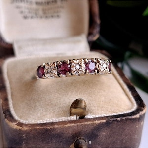 Könnte beinhalten: Ein goldener Bandring mit einem abwechselnden Muster aus drei runden roten Rubinen und zwei kleinen klaren Diamanten in quadratischen Metallfassungen. Der Ring wird in einer cremefarbenen Schmuckschatulle aus Samt präsentiert.