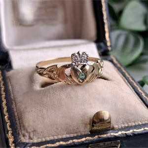 May include: A gold Claddagh ring featuring two hands holding a heart topped with a crown. The heart is set with a small green gemstone, and the crown is accented with tiny clear stones. The band displays intricate Celtic knotwork detailing.