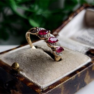 Puede incluir: Un anillo de oro con tres rubíes de corte ovalado y pequeños diamantes. El anillo está engastado en un estilo vintage y se presenta en un joyero forrado de terciopelo. El diseño del anillo es elegante y clásico.