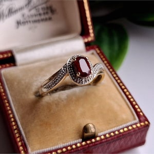 Könnte beinhalten: Ein goldener Ring mit einem großen ovalen Rubin-Mittelstein, umgeben von kleinen Diamanten. Der Ring ist in einem goldenen Band mit zusätzlichen Diamantakzenten gefasst. Der Ring wird in einer rot-goldenen Schmuckschatulle mit cremefarbenem Innenraum präsentiert.