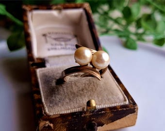May include: A vintage gold-tone ring with two creamy pearls. The ring is displayed in an antique, patterned jewelry box with a velvet interior. The ring's design features a flowing, asymmetrical shape, creating an elegant and classic look.