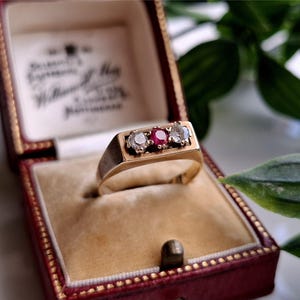 May include: A gold ring with three stones: a central red stone flanked by two clear stones. The ring is displayed in a red jewelry box with a cream-colored interior. The box is open, revealing the ring.