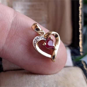Può includere: Un ciondolo a forma di cuore dorato con una gemma rossa al centro e piccole gemme bianche. Il ciondolo è tenuto tra le dita, mostrando il suo design e il suo colore.