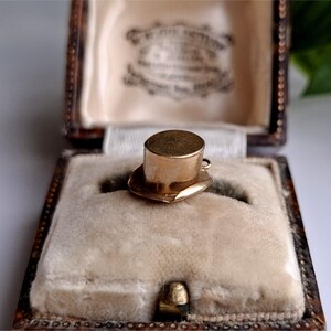 May include: A gold-coloured miniature top hat charm, set on a heart-shaped base, inside a vintage jewellery box. The box is lined with cream-coloured velvet and has a decorative, aged appearance. The charm is likely a pendant or a small collectible item.
