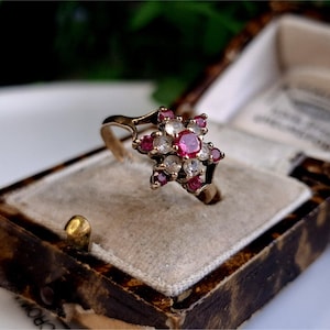 May include: A gold ring with a star-shaped cluster of red and white gemstones. The ring is in a small, brown velvet box.