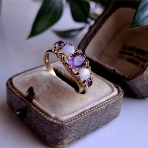 May include: A gold ring with an ornate filigree design, set with oval purple amethyst and round white opal gemstones. The ring is displayed in a vintage, brown jewelry box with a velvet interior. The ring is a size 7.