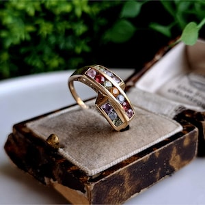Könnte beinhalten: Ein goldener Ring mit einem Band aus mehrfarbigen Edelsteinen. Der Ring liegt in einer Samt-Ringbox.