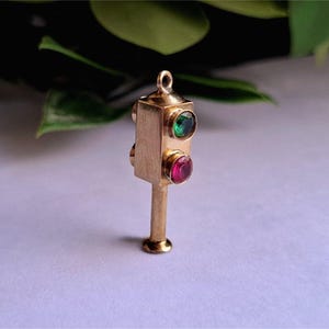 May include: A gold-colored traffic light charm with a green and a red gemstone. The charm has a small loop at the top for attaching to a necklace or bracelet. The charm is on a white surface with green leaves in the background.