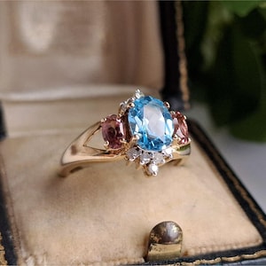 Puede incluir: Un anillo de oro con una gran gema azul ovalada, flanqueada por dos gemas rosas más pequeñas. El anillo está engastado con pequeñas piedras blancas y se exhibe en un joyero vintage.